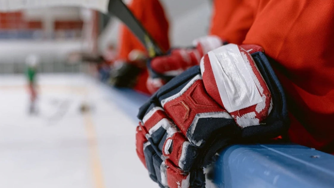 Photo of a hockey player standing in the bench area of an ice rink, wearing navy, red and white gloves