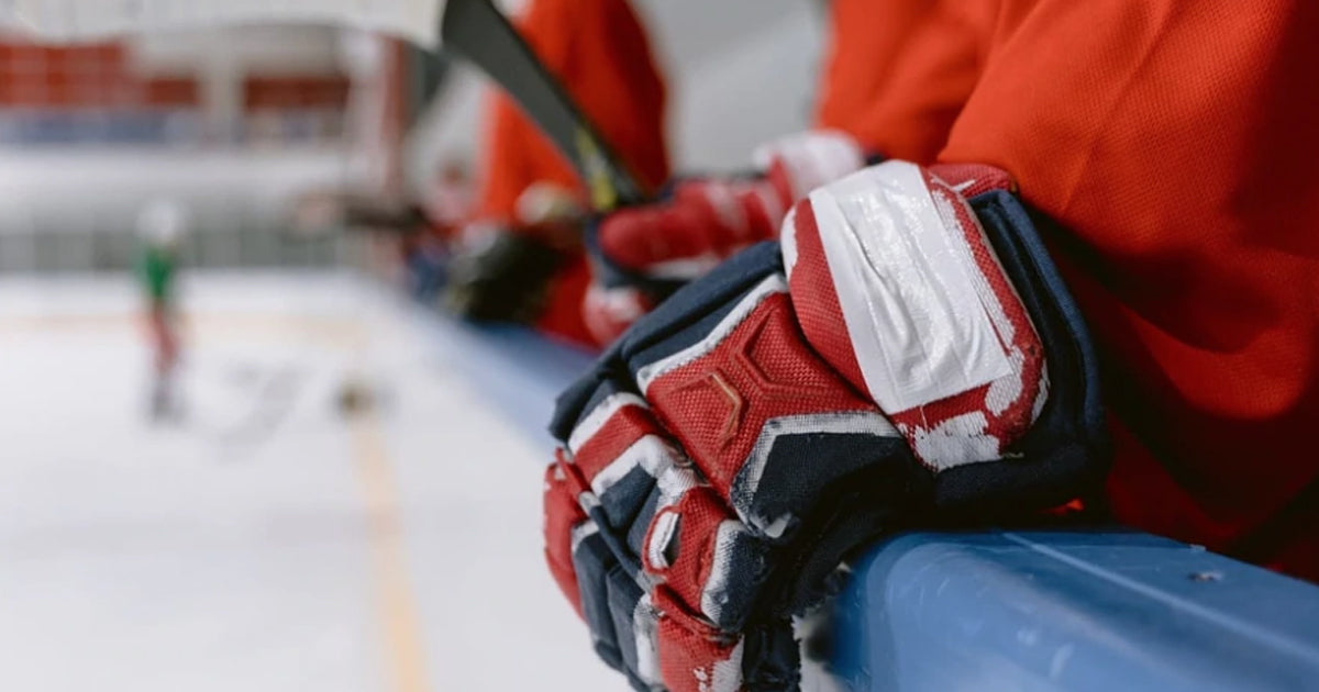 Photo of a hockey player standing in the bench area of an ice rink, wearing navy, red and white gloves 